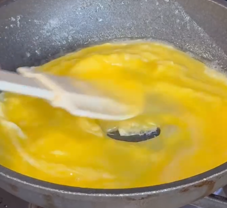 A close-up of eggs being stirred with a spatula in a frying pan on a stove, as they cook and begin to form soft scrambled eggs.