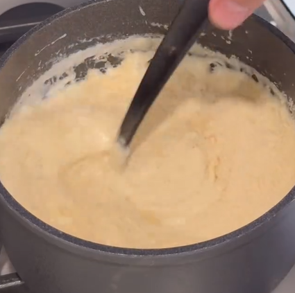 A hand stirs a thick, creamy mixture in a black saucepan on a stovetop with a black utensil.