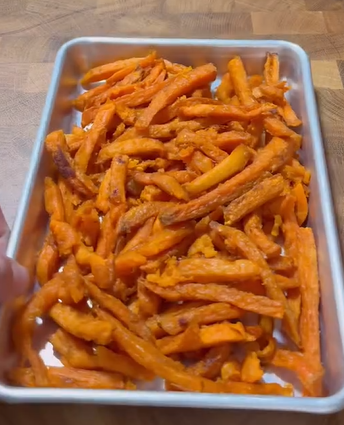 A metal tray filled with crispy, golden-orange sweet potato fries sits on a wooden surface.