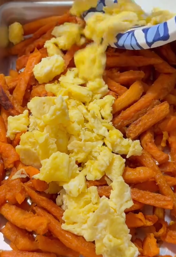 Scrambled eggs are being served onto a tray filled with sweet potato fries.