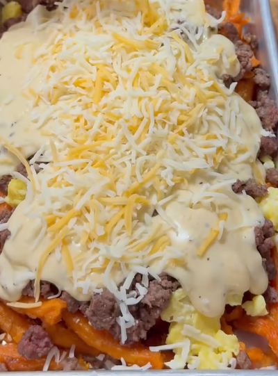 A close-up of a dish topped with shredded cheese, creamy sauce, ground beef, and scrambled eggs on a bed of curly fries. The ingredients are layered on a baking tray.