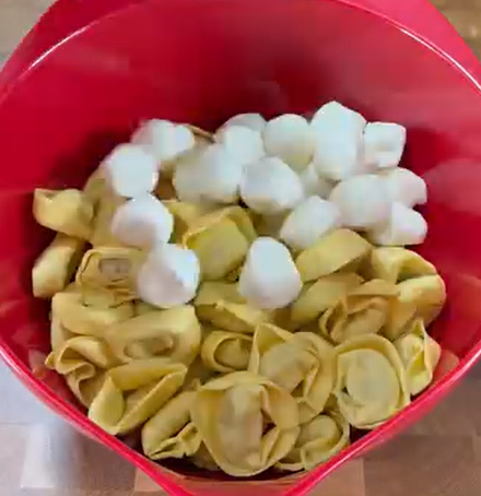 A red mixing bowl containing uncooked cheese tortellini pasta and a pile of mini mozzarella balls on a wooden surface.