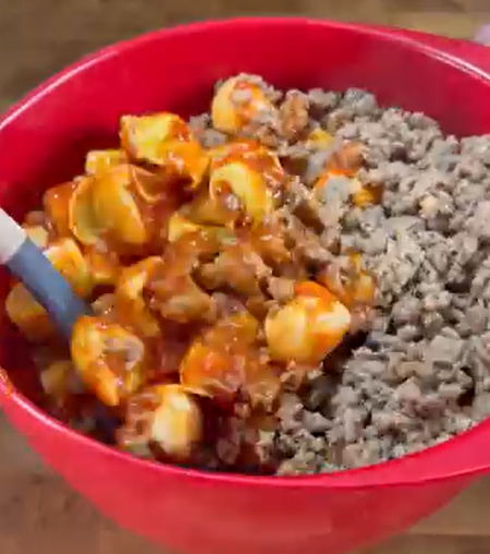 A red mixing bowl containing cooked ground beef and cheese tortellini mixed with tomato sauce, with a blue spatula resting inside.