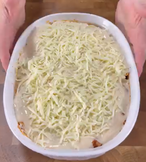 A pair of hands holds a white oval baking dish filled with an unbaked casserole topped with grated cheese and a creamy sauce, set on a wooden surface.