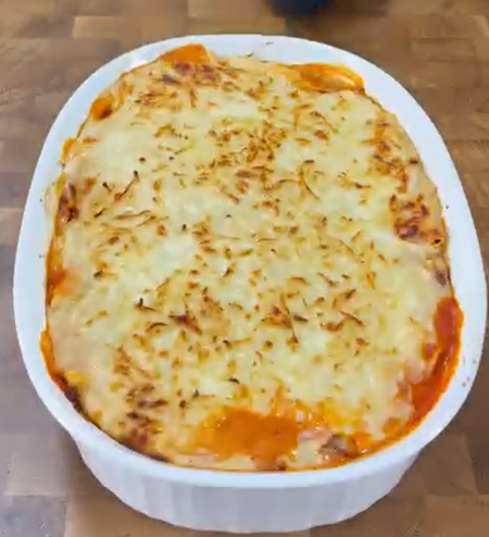 A baked lasagna sits in a white oval dish on a wooden surface, topped with melted and slightly browned cheese.