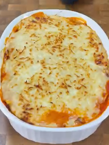 A freshly baked lasagna in a white oval dish sits on a wooden surface, topped with a golden-brown layer of melted cheese and slightly browned edges.