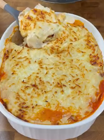 A baked pasta dish topped with melted, golden-brown cheese is shown in a white casserole dish. A serving is being lifted out, revealing layers of sauce and pasta inside.