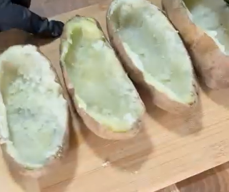 Four hollowed-out baked potato halves are lined up on a wooden cutting board, ready to be filled. A gloved hand is visible near the top left corner.