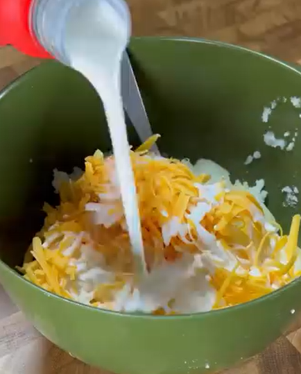 A green bowl filled with shredded yellow cheese and other ingredients as milk is being poured in, with a spoon visible inside the bowl.