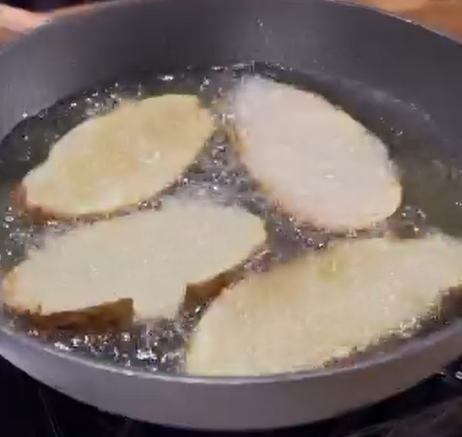 Four slices of bread frying in hot oil in a black pan on a stovetop, with bubbles forming around the bread.