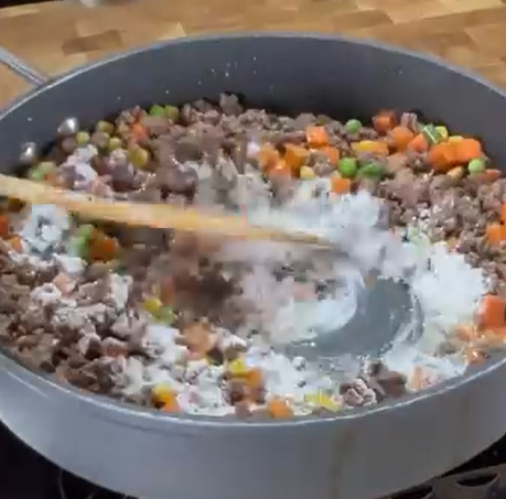 A close-up of a pan on a stove filled with ground beef, diced carrots, peas, and a white powder being stirred in with a wooden spoon. The background shows a wooden countertop.