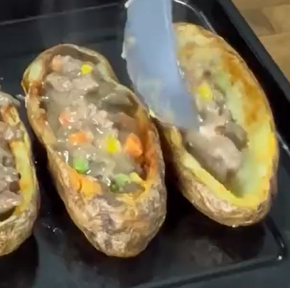 A baking tray with potato skins being filled with a mixture of meat, peas, carrots, and gravy using a large spoon. Steam is visible, indicating the food is hot.