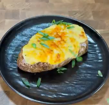 A baked potato topped with melted cheese and garnished with chopped green onions is served on a black plate, set on a wooden surface.
