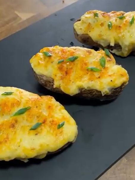 Three twice-baked potatoes topped with melted cheese and garnished with green herbs are arranged on a black slate board.
