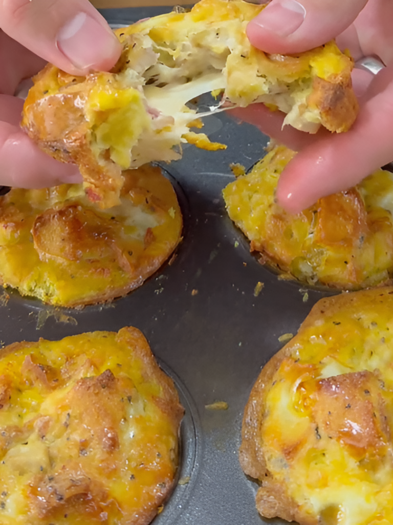 Close-up of hands pulling apart a cheesy, golden-brown muffin above a muffin pan with similar baked savory muffins, showing the gooey, melted cheese inside.