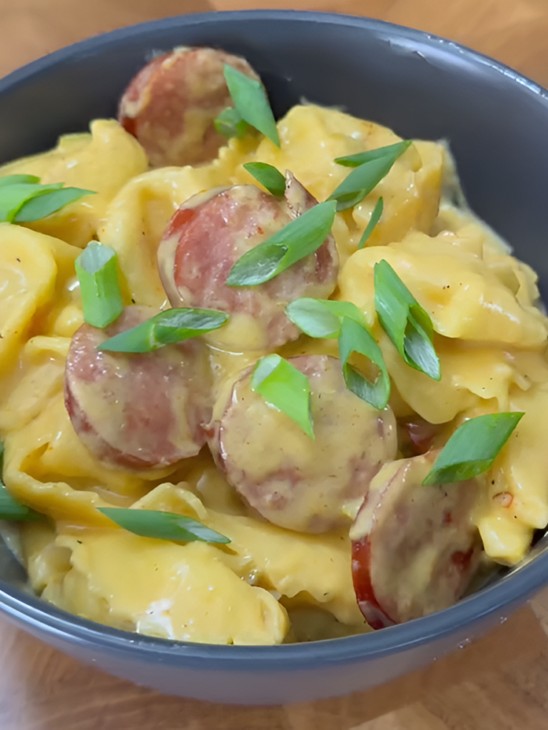 A bowl of creamy tortellini pasta with sliced sausage and a rich yellow cheese sauce, topped with fresh green onion slices.