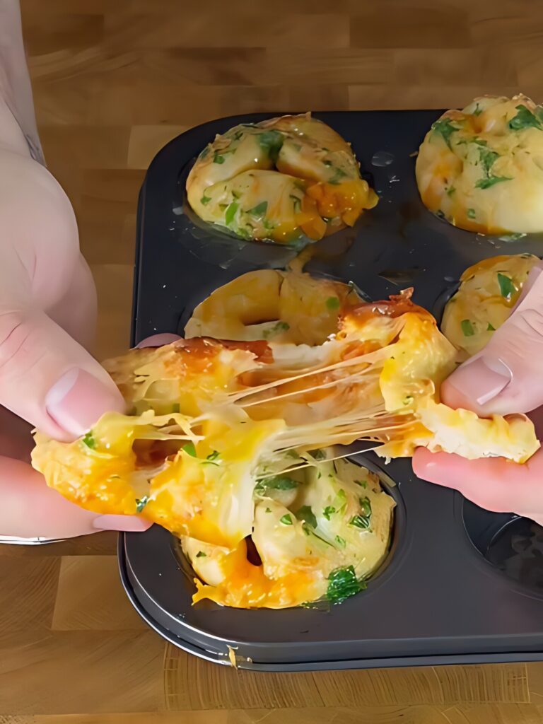 A person pulls apart a cheesy biscuit, showing melted cheese stretching between the halves, with more baked biscuits in a muffin tin in the background.