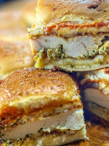 Close-up of stacked breaded chicken sandwiches, layered with mozzarella cheese, marinara sauce, and pesto, on toasted bread. The golden crust and juicy chicken are visible in the cross-section.