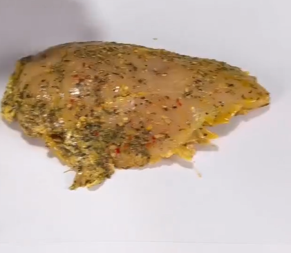 A seasoned raw chicken breast with visible herbs and spices rests on a plain white surface.