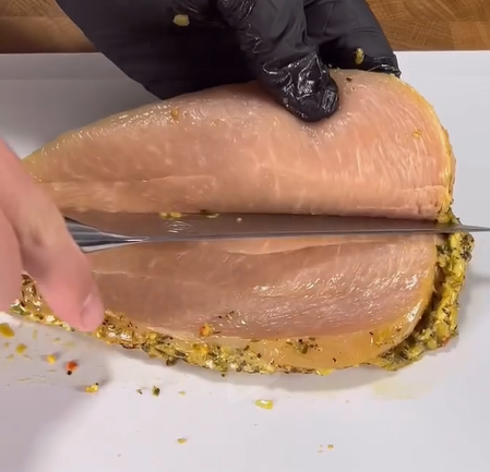 A person wearing a black glove slices through a seasoned, raw chicken breast placed on a white cutting board.
