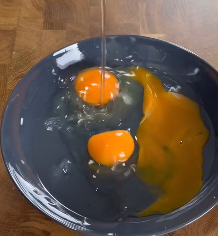 A close-up of two raw eggs cracked into a dark bowl, with one yolk intact and the other yolk broken and spreading across the bowl.