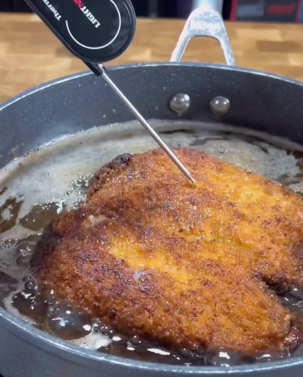 A breaded cutlet is being fried in a pan of hot oil, with a food thermometer inserted to check its internal temperature. The cutlet appears golden brown and crispy.