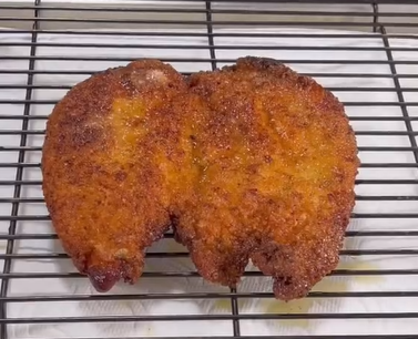 A large, golden-brown breaded and fried cutlet rests on a metal cooling rack with a paper towel underneath. The surface appears crispy and oily, and the cutlet has an uneven, slightly irregular shape.