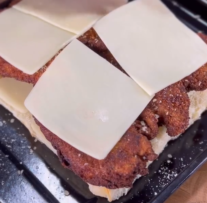 A close-up of a sandwich with crispy fried chicken pieces and square slices of cheese on white bread, served on a black tray.
