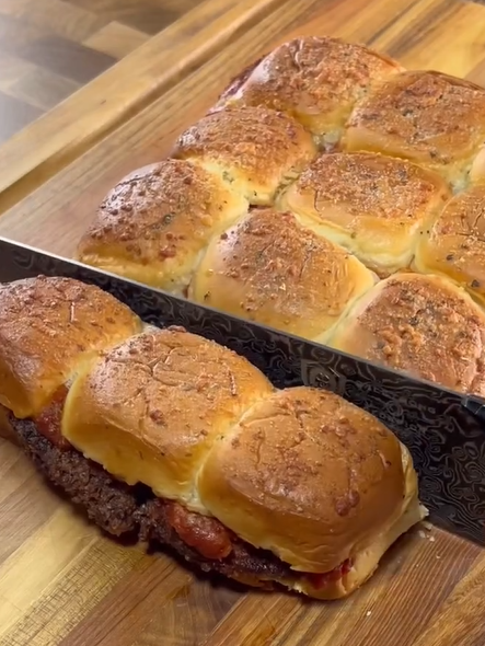 A large knife slices through a batch of baked slider sandwiches filled with hamburger patties and sauce on a wooden cutting board.