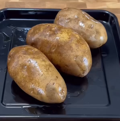 Three whole, unpeeled potatoes sit on a black baking tray, lightly coated with water or oil. The tray is on a wood-patterned surface.