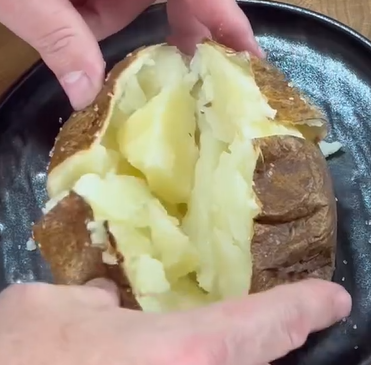 A close-up of hands breaking open a baked potato on a black plate, revealing its fluffy, steamy interior.