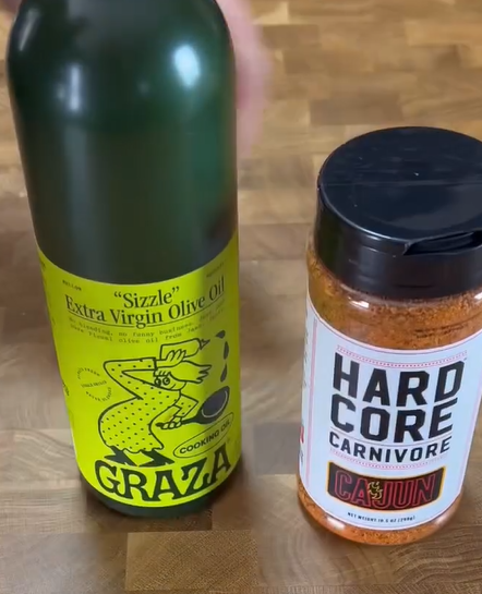A bottle of Graza Sizzle extra virgin olive oil with a bright green label sits next to a container of Hard Core Carnivore Cajun seasoning on a wooden surface.