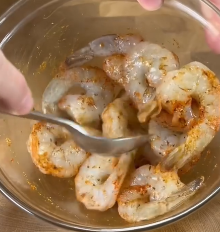 A close-up of raw shrimp being mixed with spices in a metal bowl using a spoon.