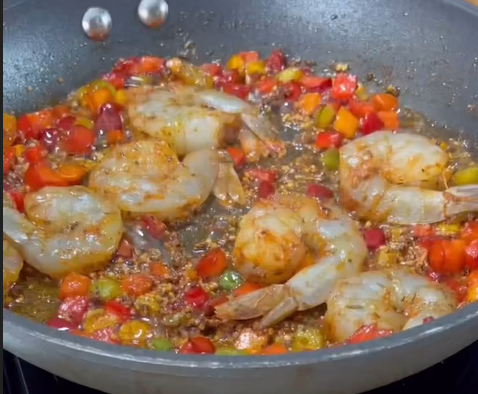 Large shrimp are being sautéed in a pan with diced red, yellow, and green bell peppers and spices, creating a colorful and flavorful mixture.