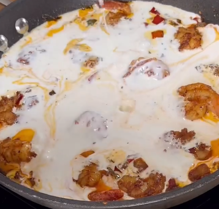 A skillet filled with shrimp simmering in a creamy white sauce, with visible pieces of red and yellow bell peppers and seasonings.