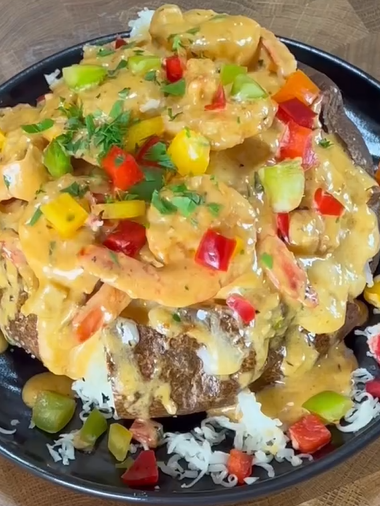 A baked potato topped with creamy shrimp, colorful diced bell peppers, and fresh herbs, served on a black plate with shredded cheese garnished around the base.