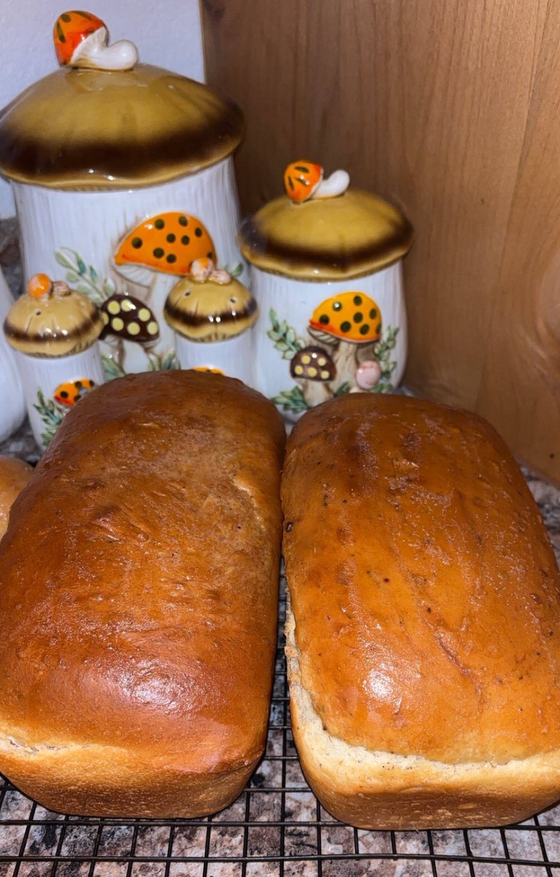 Two golden-brown loaves of bread cool on a wire rack in front of ceramic kitchen canisters decorated with orange mushroom lids and small painted mushrooms, set against a wooden backdrop.