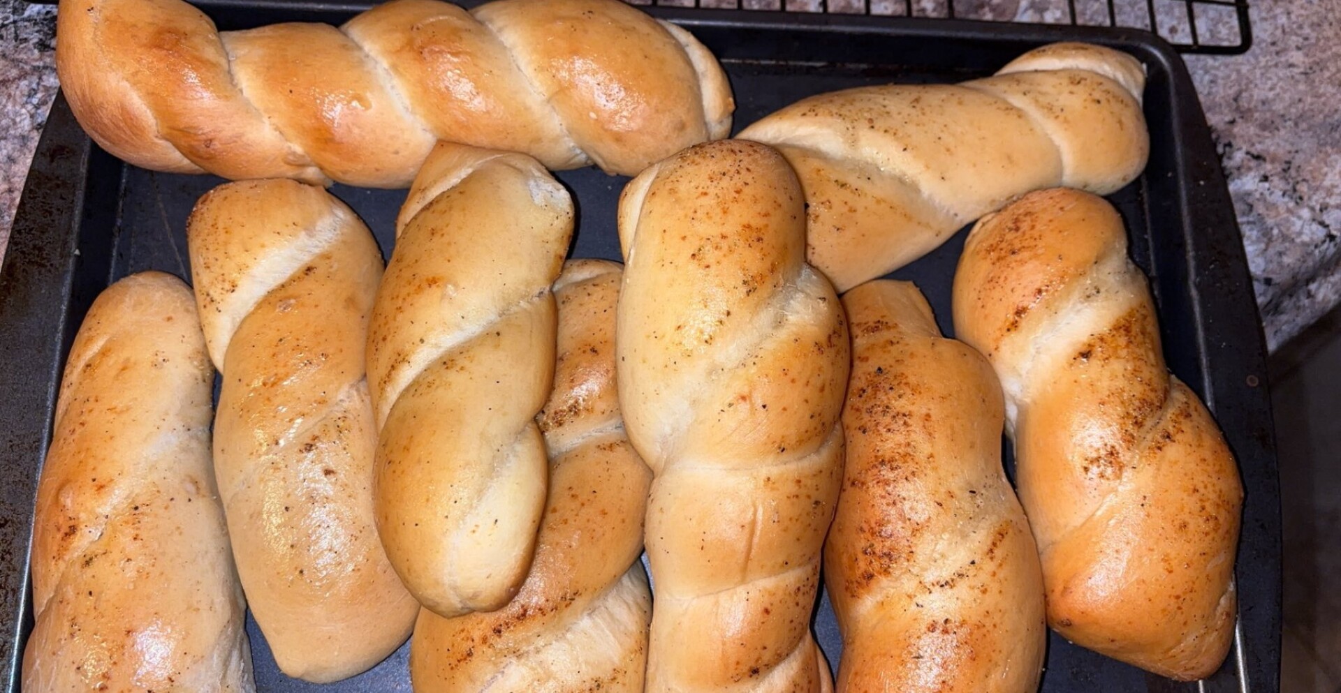 A baking tray with nine freshly baked, golden-brown twisted breadsticks, some sprinkled lightly with seasoning, resting on a countertop.