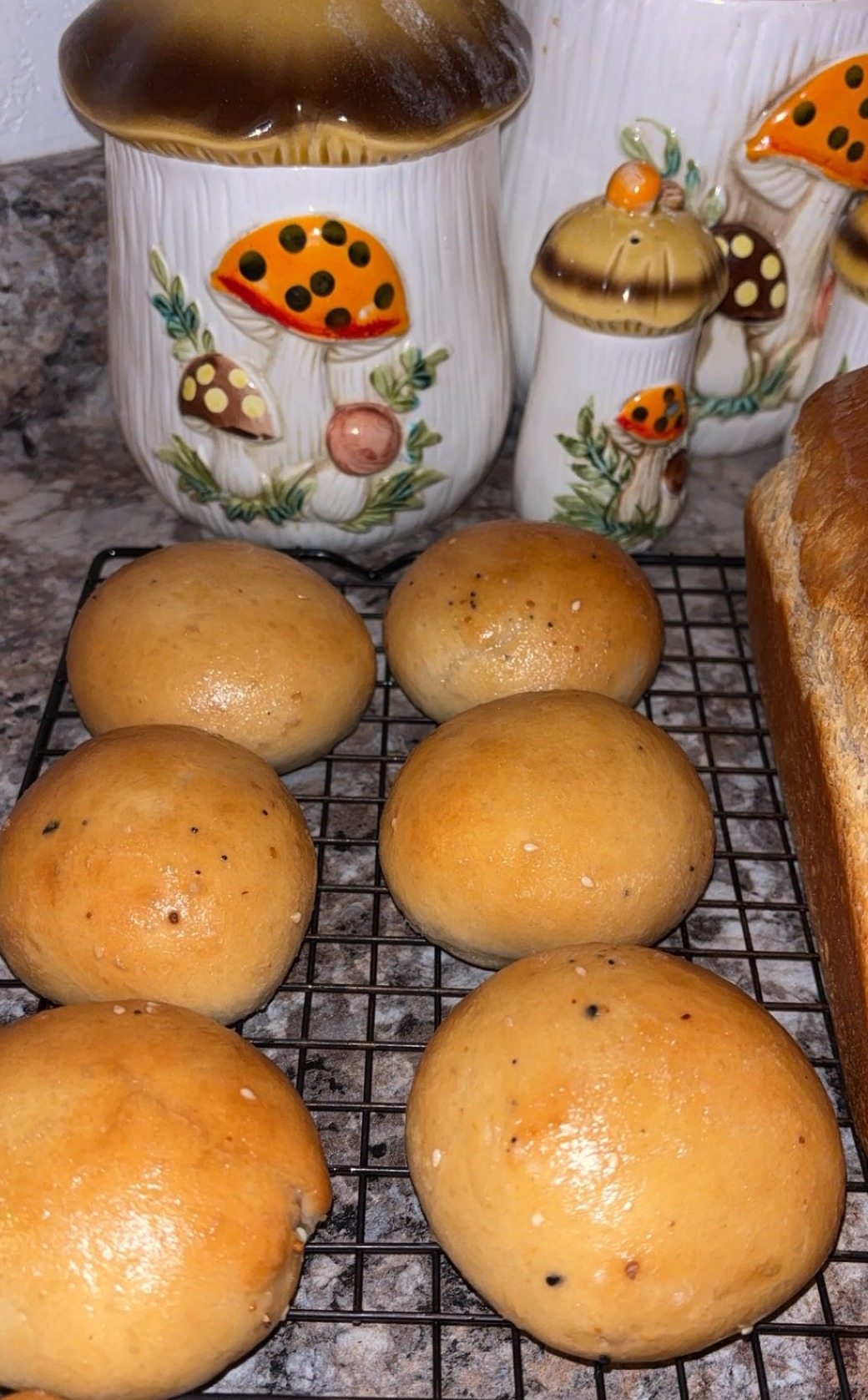 Six golden brown bread rolls are cooling on a wire rack next to a loaf of bread. In the background, ceramic kitchen canisters decorated with colorful mushroom designs are visible on a countertop.