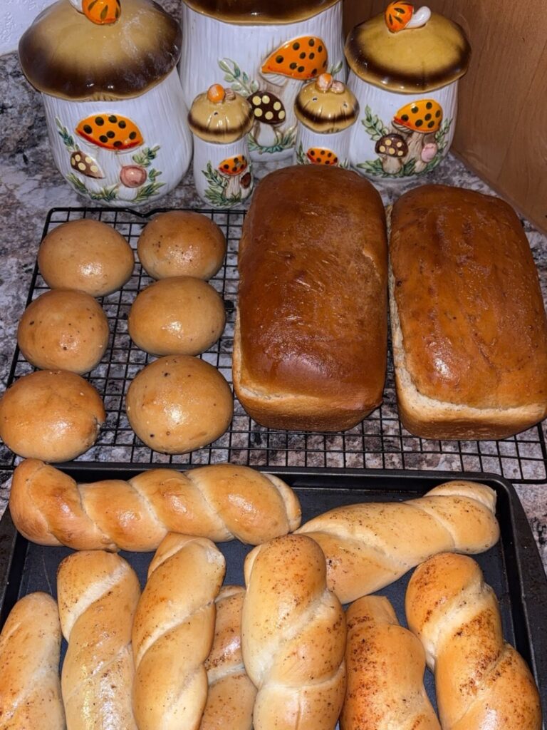 A variety of freshly baked bread, including round rolls, two loaves, and twisted breadsticks, cooling on racks and a tray. Behind them are decorative ceramic jars with mushroom and ladybug designs.