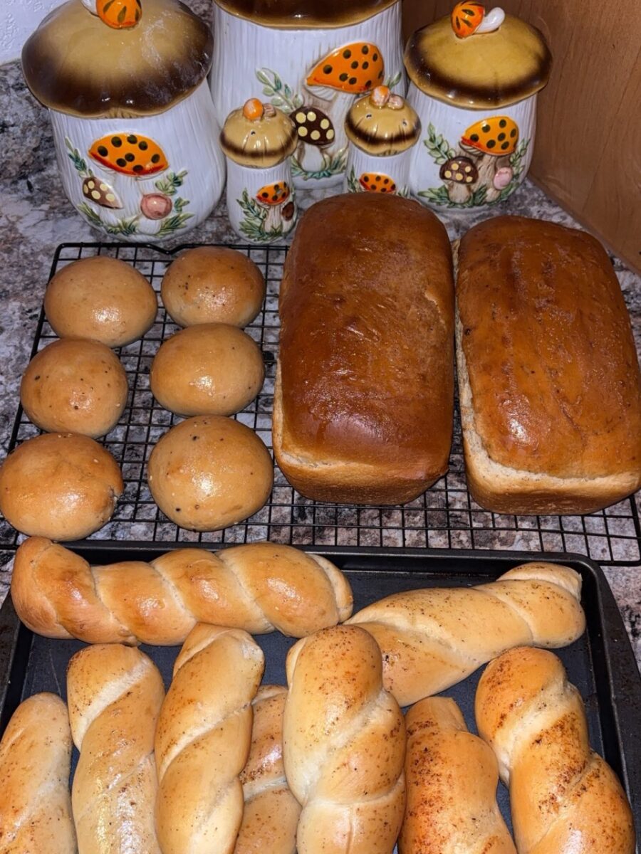 A variety of freshly baked bread, including round rolls, two loaves, and twisted breadsticks, cooling on racks and a tray. Behind them are decorative ceramic jars with mushroom and ladybug designs.