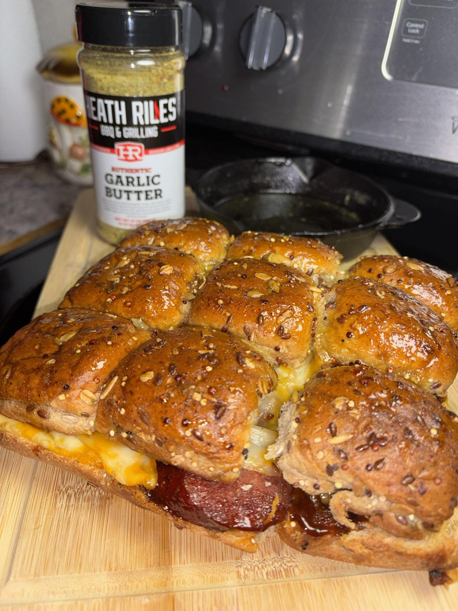 A cheesy pull-apart sandwich with pepperoni and toasted seeded buns sits on a wooden cutting board. Behind it, there is a jar of Death Riles Garlic Butter seasoning and a small skillet on a stovetop.
