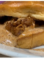 Close-up of a fried chicken sandwich with a glossy bun and creamy, pepper-speckled sauce spilling out onto a plate.