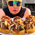 A person wearing reflective sunglasses holds a plate of sandwiches topped with shredded slaw and sauce, with an outdoor grill in the background.