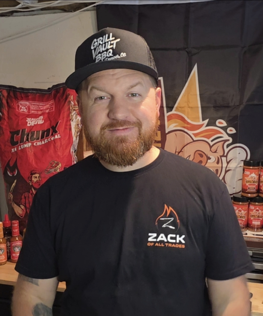A bearded man wearing a Grill Vault BBQ cap and a Zack of All Trades t-shirt smiles in front of barbecue-themed decor, including bags of charcoal and hot sauce bottles.