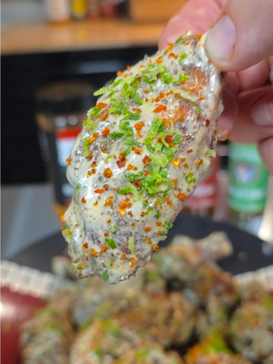 A close-up of a hand holding a seasoned chicken wing coated in creamy sauce, garnished with red pepper flakes and green herbs, with more wings blurred in the background.