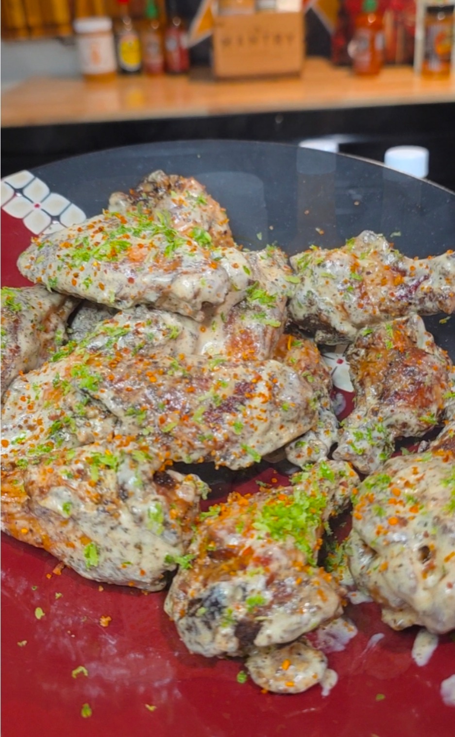 A plate of creamy, seasoned chicken wings garnished with chopped herbs and spices, served on a red dish with a kitchen counter and various sauce bottles in the background.
