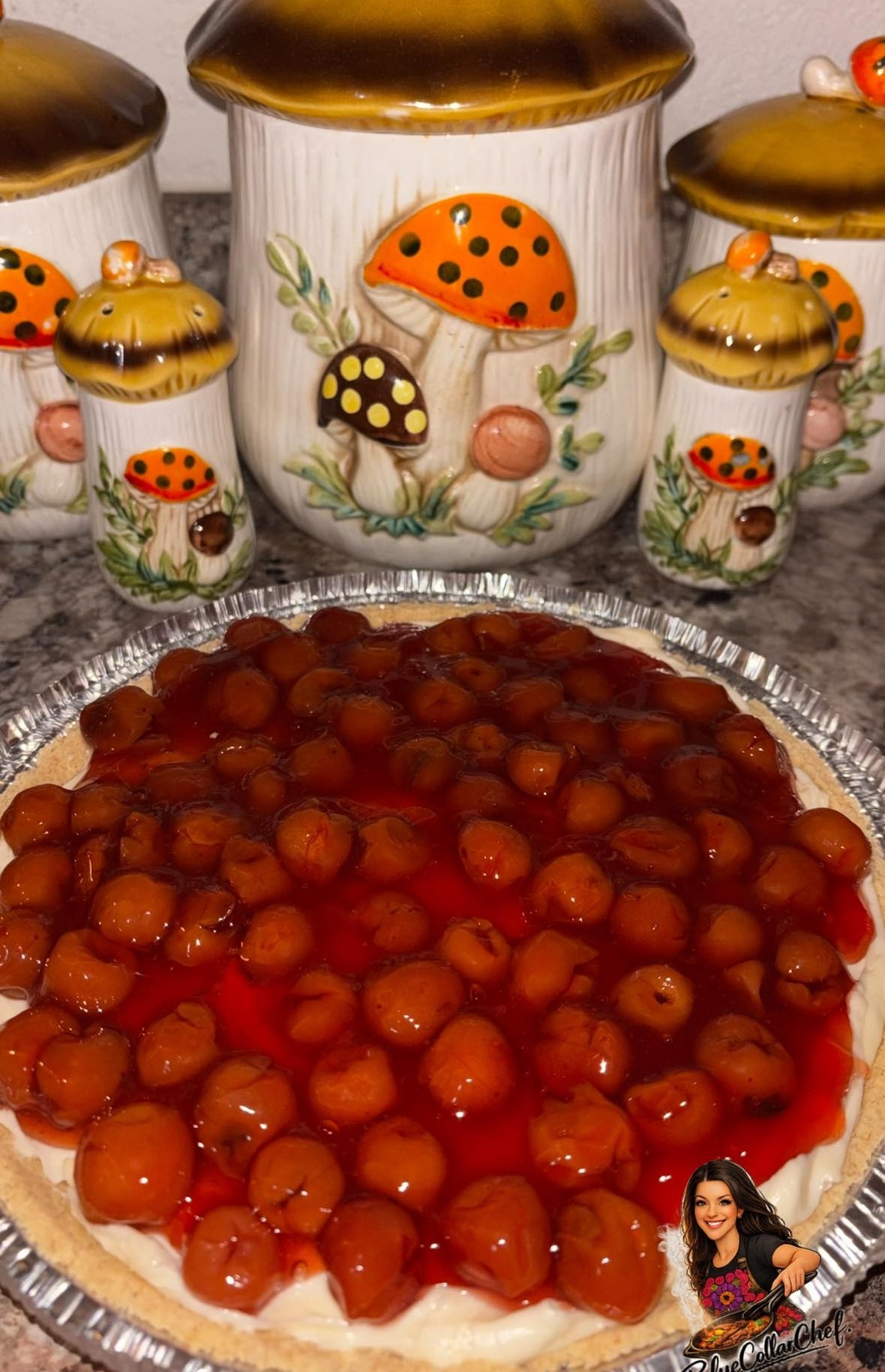 A cherry cheesecake in a foil pie tin sits on a kitchen counter. Behind it are four ceramic canisters decorated with orange and yellow mushrooms and green leaves.