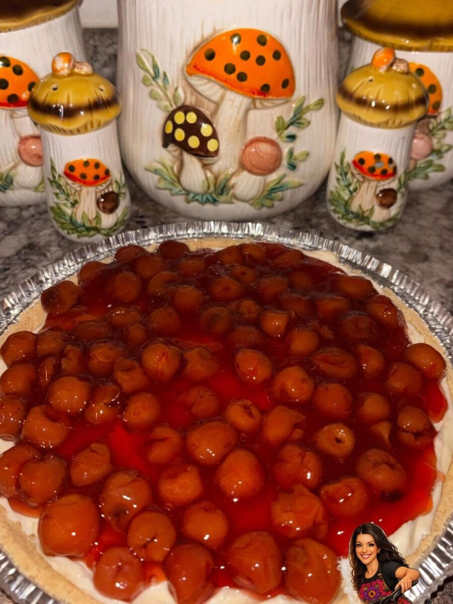 A cherry-topped dessert pie sits on a counter in front of vintage mushroom-themed ceramic canisters. There is a small sticker of a smiling woman in the bottom right corner.