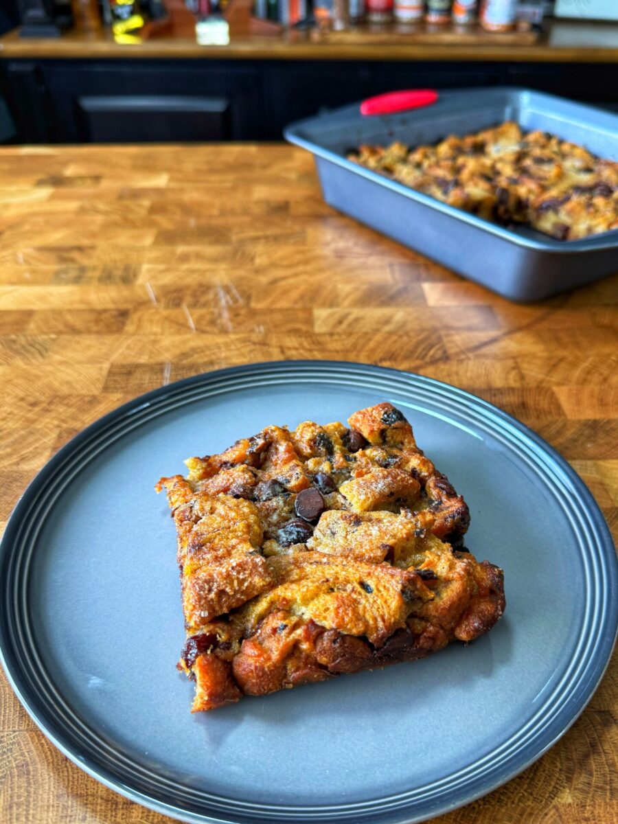 A square piece of bread pudding with chocolate chips sits on a gray plate, with the rest of the dessert visible in a baking pan on a wooden countertop in the background.